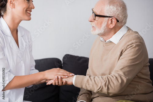 side view of aged man in eyeglasses holding hands with social worker at home
