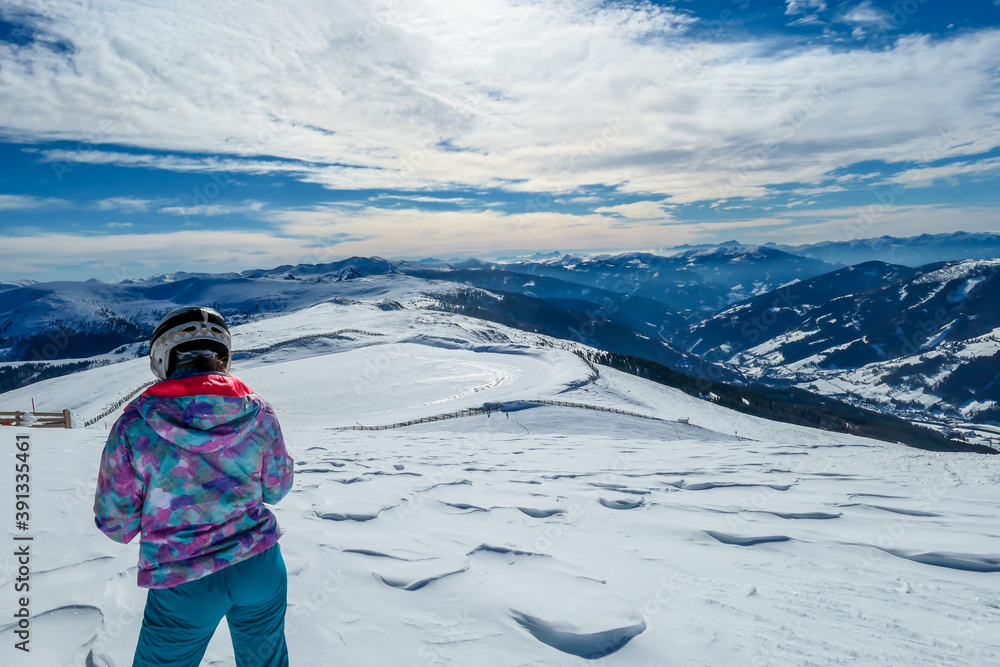 A woman standing on the snow on top of Katschberg in Austria. She is happy and joyful. Panoramic view on the surrounding mountains. Winter wonderland. Bright and sunny winter day. Love and happiness