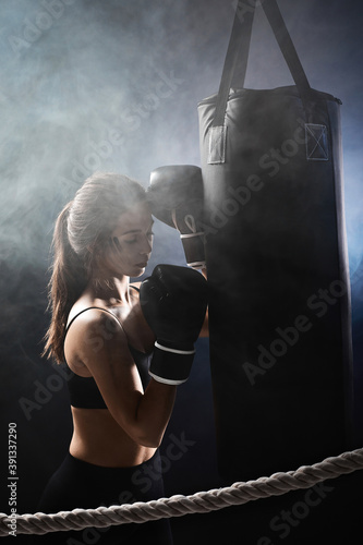 athletic woman in black boxing gloves leaning on punching bag on dark studio background. woman resting after workout © producer