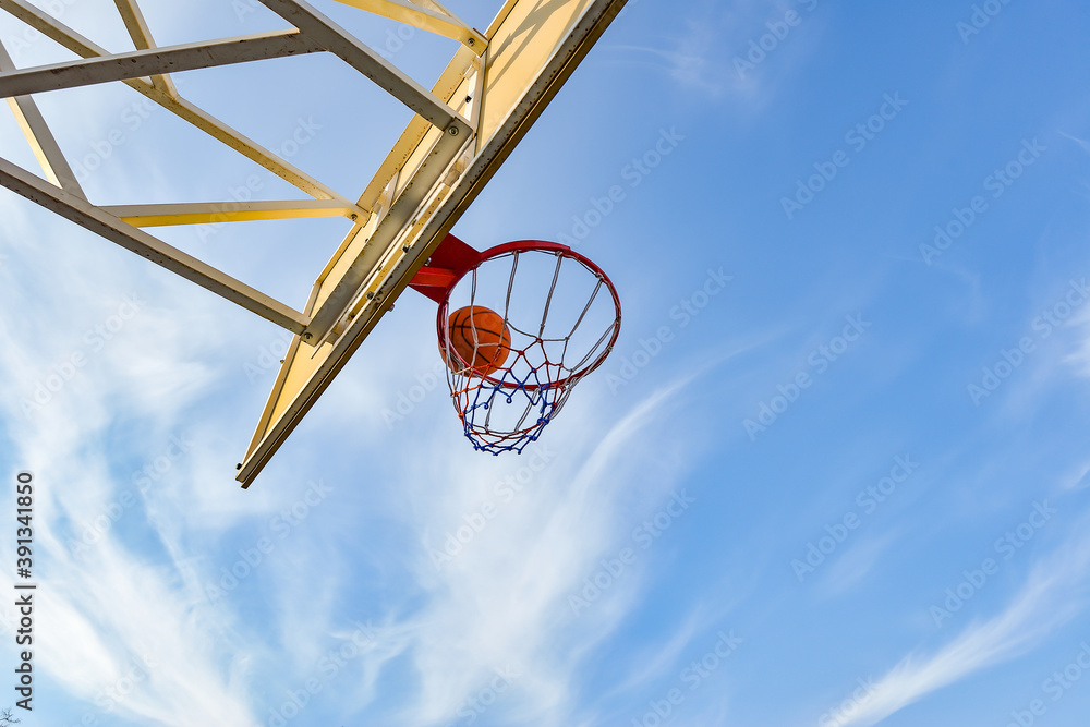 A basketball ball flies into a hoop with a net against a blue cloudy sky. Sports activities on the playground.