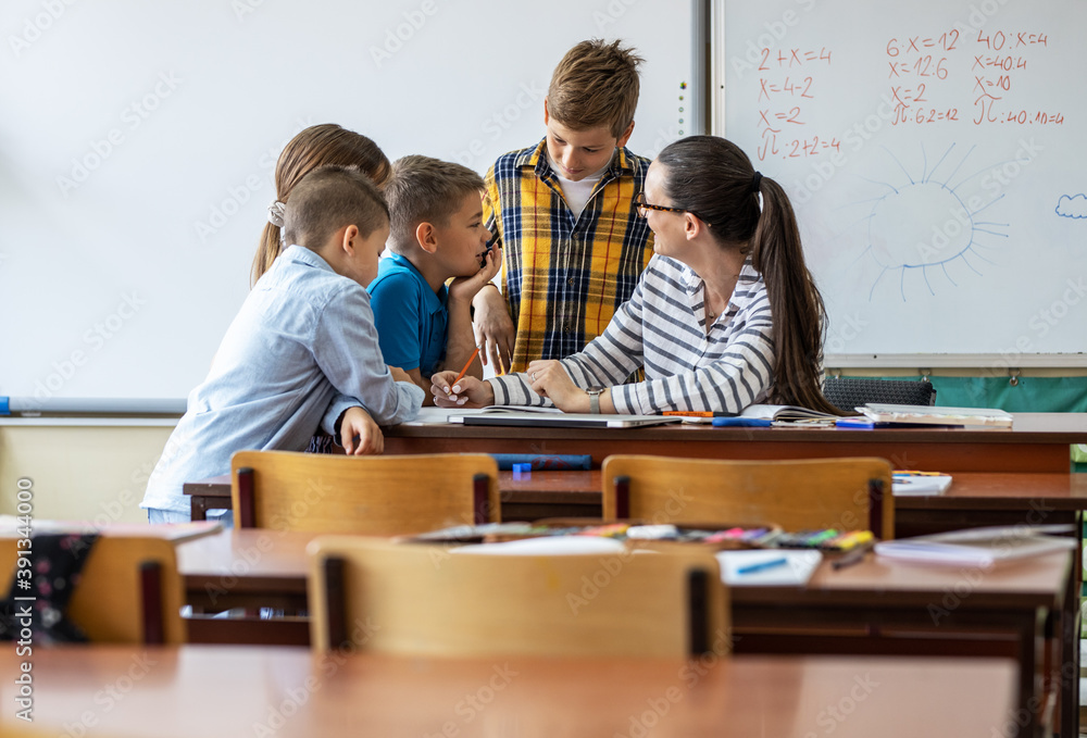 Female teacher helps school kids to finish they lesson.They sitting all together at one desk.	
