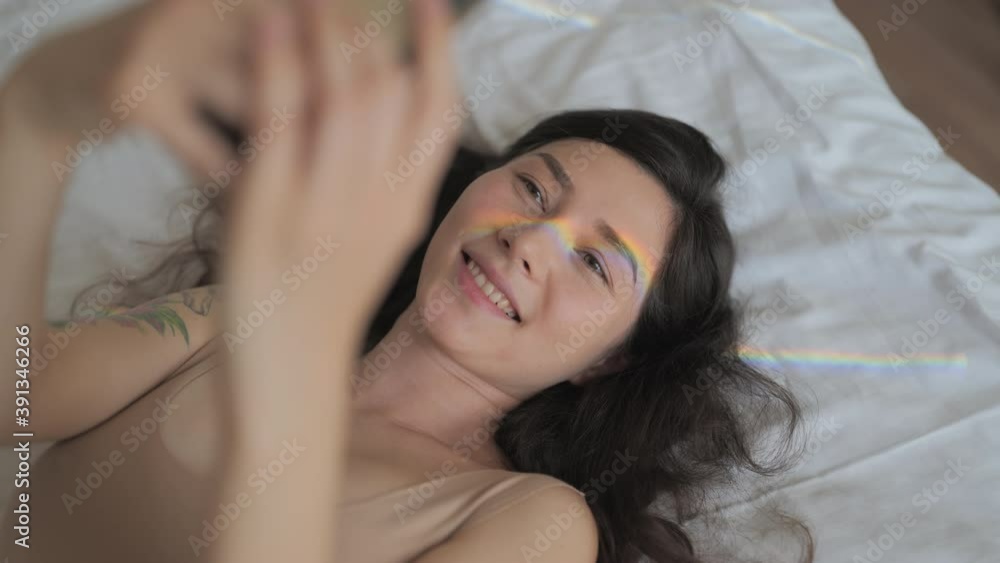 A happy mixed race woman is taking selfie photo with rainbow flare on her face on her smartphone while lying on bed in sunny room. happiness, lgbt, lesbian concept. A happy mixed race woman is taking selfie photo with rainbow flare on her face on her smartphone while lying on bed in sunny room. happiness, lgbt, lesbian concept.