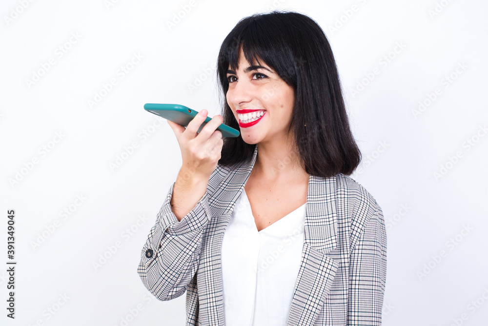 Caucasian woman standing against white background wearing blazer, holding smartphone and sending voice message. Caucasian woman standing against white background wearing blazer, holding smartphone and sending voice message.