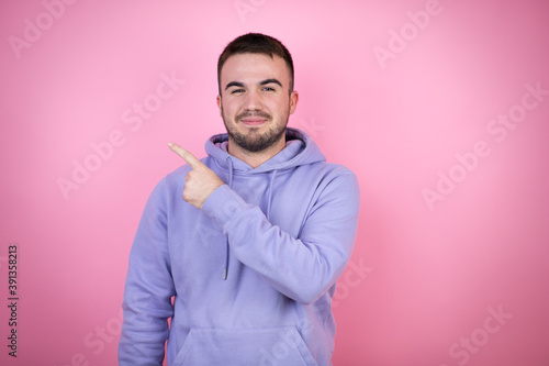 Young handsome man wearing casual sweatshirt over isolated pink background smiling and pointing with hand and finger to the side © Irene