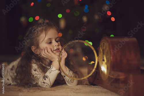 A cute girl lies on the floor with her chin resting on her palms and looks at a large wooden clock with a glass cover. Bokeh. Christmas and New Year concept