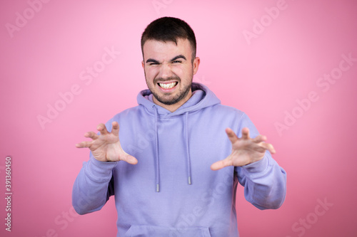 Young handsome man wearing casual sweatshirt over isolated pink background smiling funny doing claw gesture as cat, aggressive and sexy expression © Irene