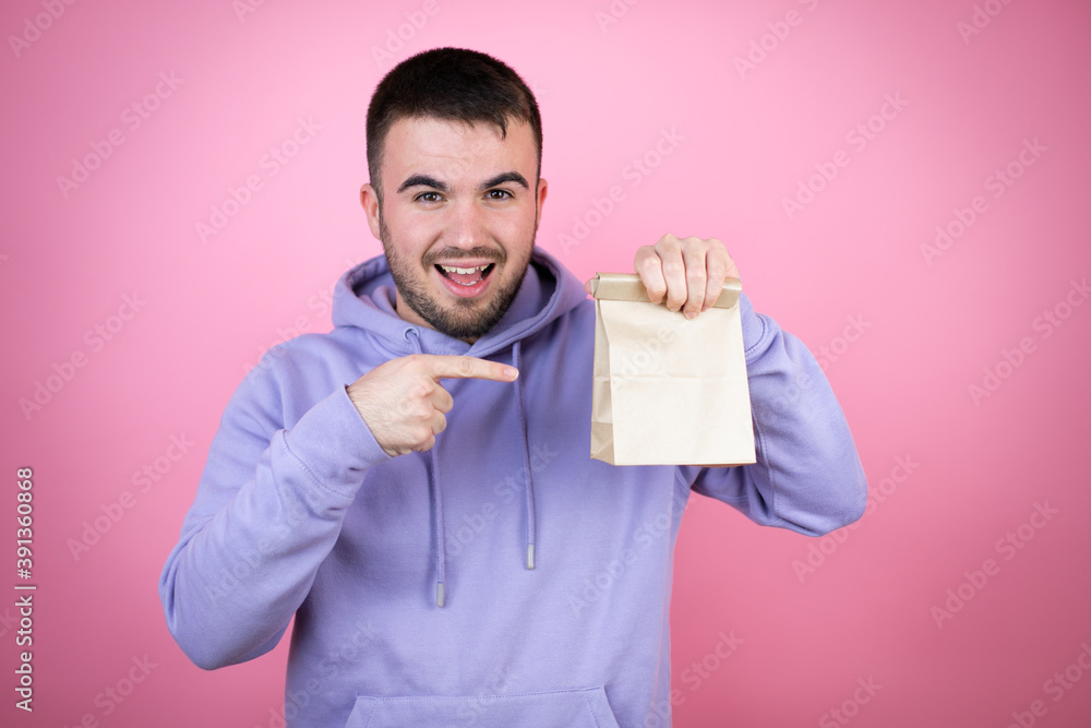 Young handsome man wearing casual sweatshirt over isolated pink background smiling, holding a paper bag and pointing the bag Young handsome man wearing casual sweatshirt over isolated pink background smiling, holding a paper bag and pointing the bag
