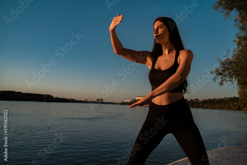 Young adult woman doing tai chi exercises by the river at sunset, wellbeing and health 
