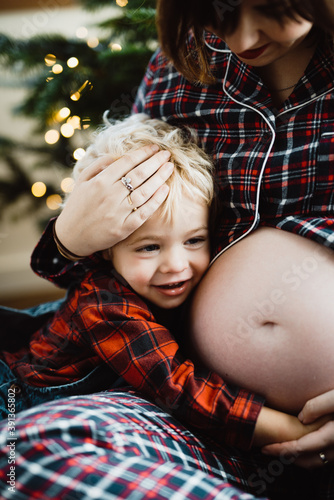 toddler (little boy) hugginh the pregnant belly of his mother wearing pyjamas in front of the christmas tree with christmas lights photo