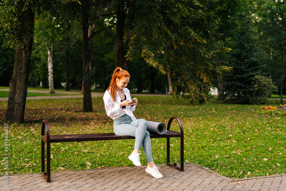 Red-haired joyful young woman wearing sportswear watching video on smartphone sitting bench at city park. Attractive female walking chatting with friends using cellphone outdoors. Red-haired joyful young woman wearing sportswear watching video on smartphone sitting bench at city park. Attractive female walking chatting with friends using cellphone outdoors.