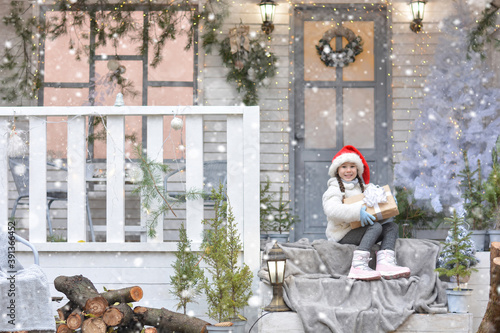 A happy girl in a fur coat, gloves, Santa hat, boots, sits on the porch of the Christmas house and holds a gift. Happy New Year and Merry Christmas