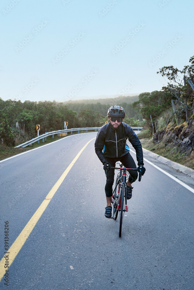 Fototapeta premium Young attractive cyclist on the road wearing black windbreaker jacket, helmet and sunglasses practicing in cycling on fresh air. Concept of healthy hobby and regular trainings, selective focu.