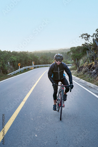 Young attractive cyclist on the road  wearing black windbreaker jacket, helmet and sunglasses practicing in cycling on fresh air. Concept of healthy hobby and regular trainings,  selective focu. © JorgeAndres
