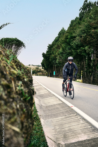 Professional male cyclist in black windbreaker jacket, protective glasses riding bicycle on paved road and green trees with blur background. Concept of summer activity and healthy lifestyle.