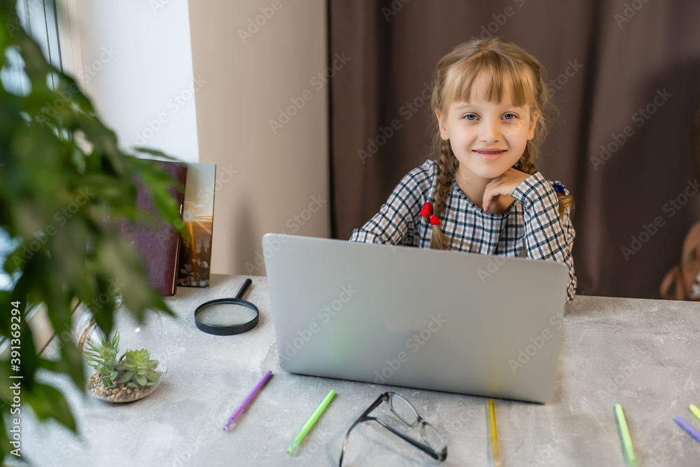 Little blonde girl doing homework at home at the table. The child is home-schooled. A girl with light hair performs a task online using a laptop and tablet computer. Little blonde girl doing homework at home at the table. The child is home-schooled. A girl with light hair performs a task online using a laptop and tablet computer.