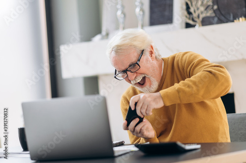 Wallpaper Mural Cheerful mature gray-haired man emotionally playing a game on his phone during work break while sitting at his workplace Torontodigital.ca