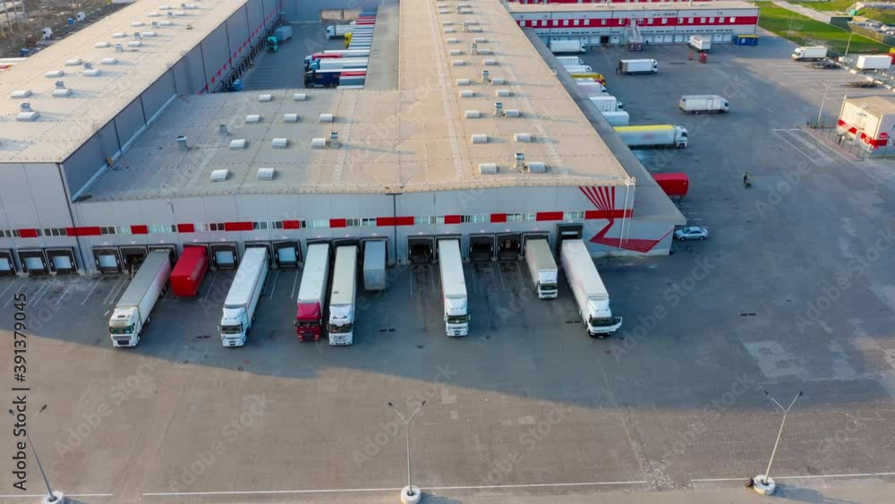 Logistics park with a warehouse - loading hub. Semi-trucks with freight trailers standing at the ramps for loading/unloading goods at sunset. Aerial hyper lapse (motion time lapse)