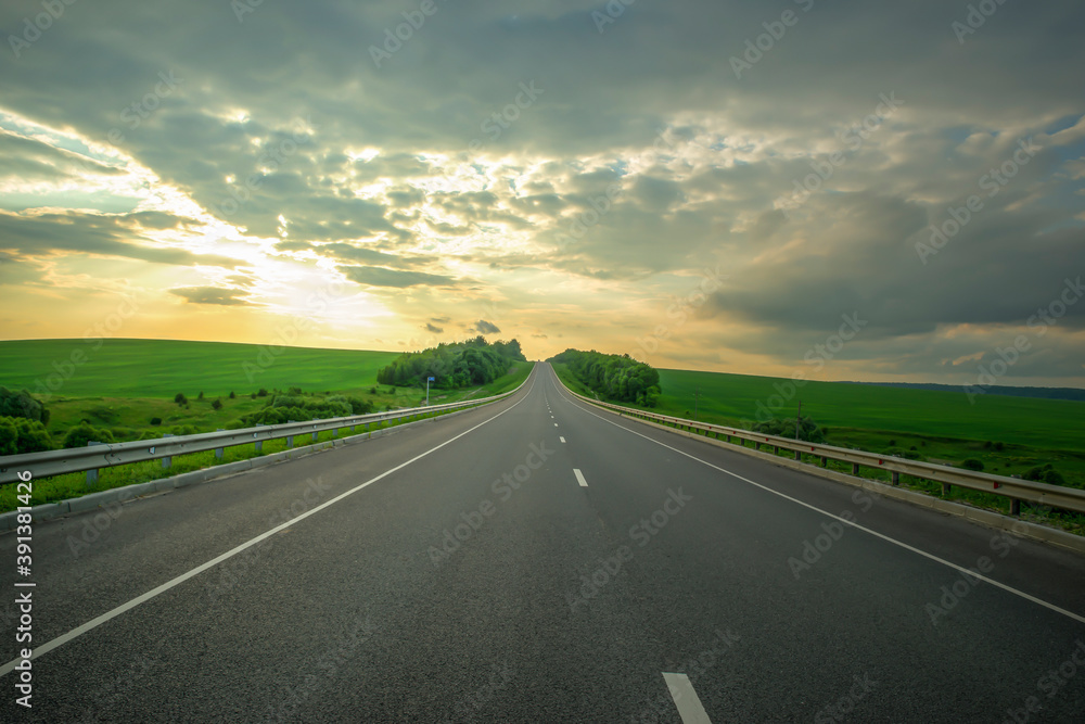 Landscape with empty asphalt road at sunset. Cloudy sky and green meadows.