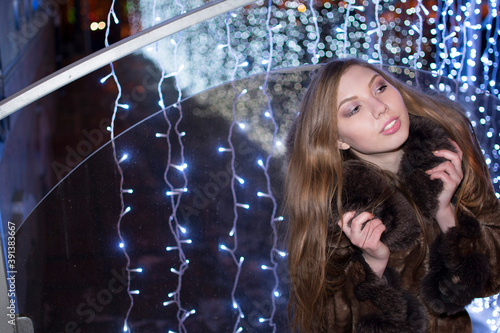 outdoor outside portrait of young luxury woman with hatless Blue bokeh blur big city street night light effect Cute flirting girl with smiley face looking at far Female holding fur collar brown coat photo