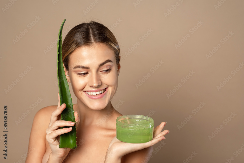 Smiling happy young female model holding aloe leaf and jar of aloe gel on beige background. The concept of skin care, moisturizing with natural cosmetics Smiling happy young female model holding aloe leaf and jar of aloe gel on beige background. The concept of skin care, moisturizing with natural cosmetics
