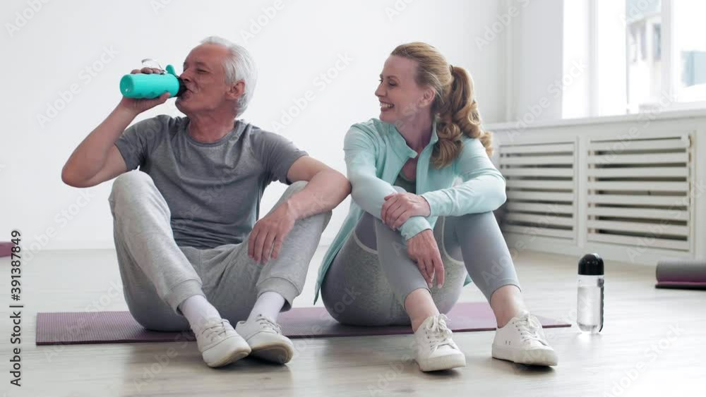 Wide shot of Caucasian senior sportsman and good-looking sportswoman wearing sportswear sitting together on mat in gym, talking and then looking at camera. Man drinking water Wide shot of Caucasian senior sportsman and good-looking sportswoman wearing sportswear sitting together on mat in gym, talking and then looking at camera. Man drinking water