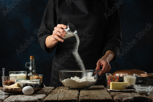 Professional chef in black uniform pours sugar into the glass bowl with flour on dark blue background. Backstage of preparing dough for pie. Frozen motion. Concept of cooking process. © Anton