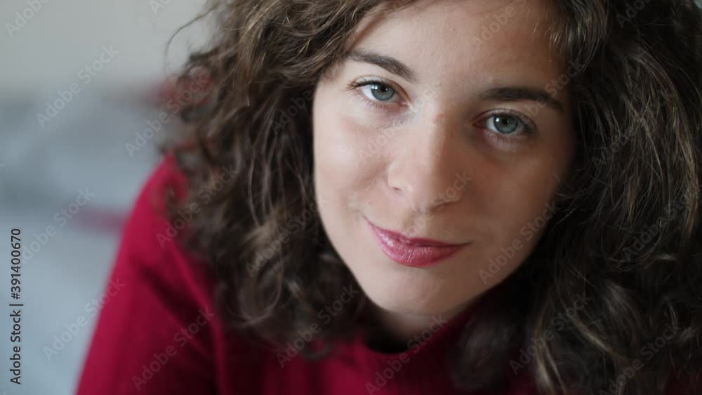 Close up of beautiful cute young woman looking up and smiling at camera. Attractive millennial girl with curly hair, red pullover and natural make-up looking at camera with blue eyes. Slow-motion. Close up of beautiful cute young woman looking up and smiling at camera. Attractive millennial girl with curly hair, red pullover and natural make-up looking at camera with blue eyes. Slow-motion.