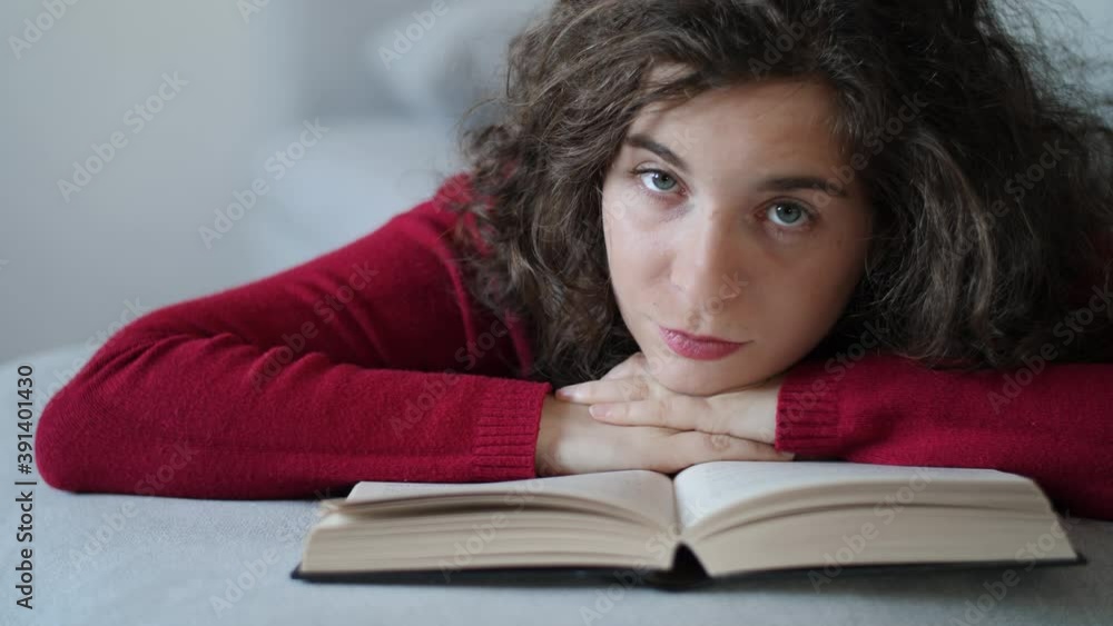Tired and bored young woman with red pullover resting her head on book. Pretty millennial girl with curly brown hair looking at camera with beautiful eyes. Slow-motion, close-up, daylight at home. Tired and bored young woman with red pullover resting her head on book. Pretty millennial girl with curly brown hair looking at camera with beautiful eyes. Slow-motion, close-up, daylight at home.
