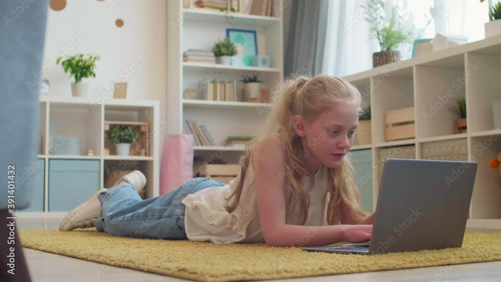 POV full shot of blond-haired Caucasian school girl laying down on carpet in her room, typing on laptop keyboard, looking up and smiling on camera POV full shot of blond-haired Caucasian school girl laying down on carpet in her room, typing on laptop keyboard, looking up and smiling on camera
