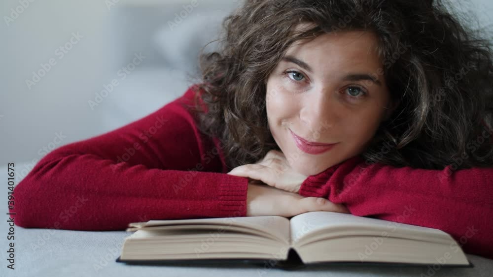 Happy young woman with red pullover resting her head on book smiling. Pretty and cute millennial girl with curly brown hair looking in love at camera with beautiful blue eyes. Slow-motion, close-up. Happy young woman with red pullover resting her head on book smiling. Pretty and cute millennial girl with curly brown hair looking in love at camera with beautiful blue eyes. Slow-motion, close-up.