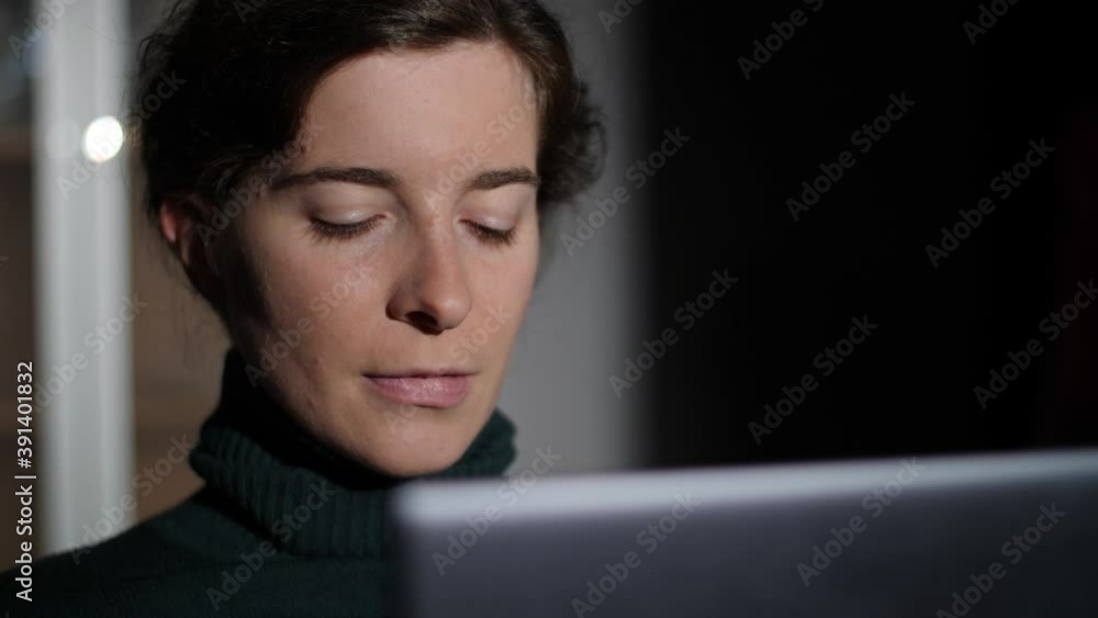 Confident young woman working with laptop at night. Stylish millennial girl writing on computer, thinking, biting lips and looking into distance. Dark background at home. Confident young woman working with laptop at night. Stylish millennial girl writing on computer, thinking, biting lips and looking into distance. Dark background at home.