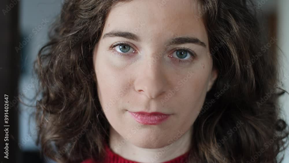 Close-up of beautiful confident young woman opening eyes and looking at camera. Attractive millennial girl with curly hair, red pullover and natural make-up at home indoors. Slow-motion. Close-up of beautiful confident young woman opening eyes and looking at camera. Attractive millennial girl with curly hair, red pullover and natural make-up at home indoors. Slow-motion.