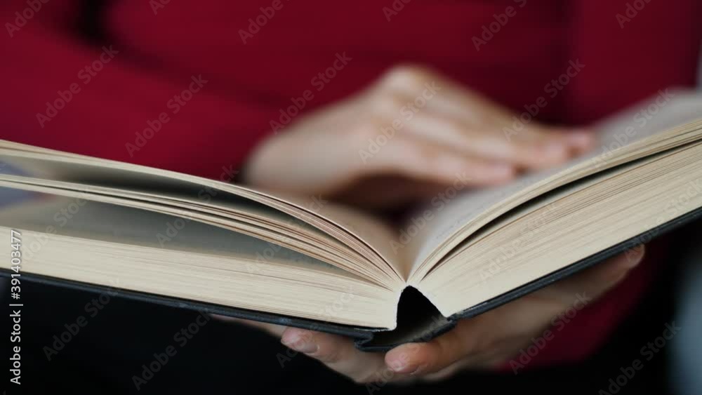 Close-up of young woman with red pullover reading hardcover book at home. Caucasian girl with pretty hands sitting while turning pages of a novel. Slow-motion, extreme close-up, natural daylight. Close-up of young woman with red pullover reading hardcover book at home. Caucasian girl with pretty hands sitting while turning pages of a novel. Slow-motion, extreme close-up, natural daylight.