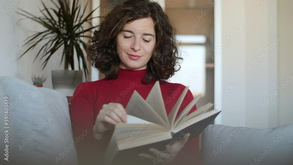 Young woman with red pullover reading hardcover book on grey couch at home. Happy beautiful girl with curly hair sitting on cozy sofa turning pages of a novel. Slow-motion, close-up, natural daylight. Young woman with red pullover reading hardcover book on grey couch at home. Happy beautiful girl with curly hair sitting on cozy sofa turning pages of a novel. Slow-motion, close-up, natural daylight.