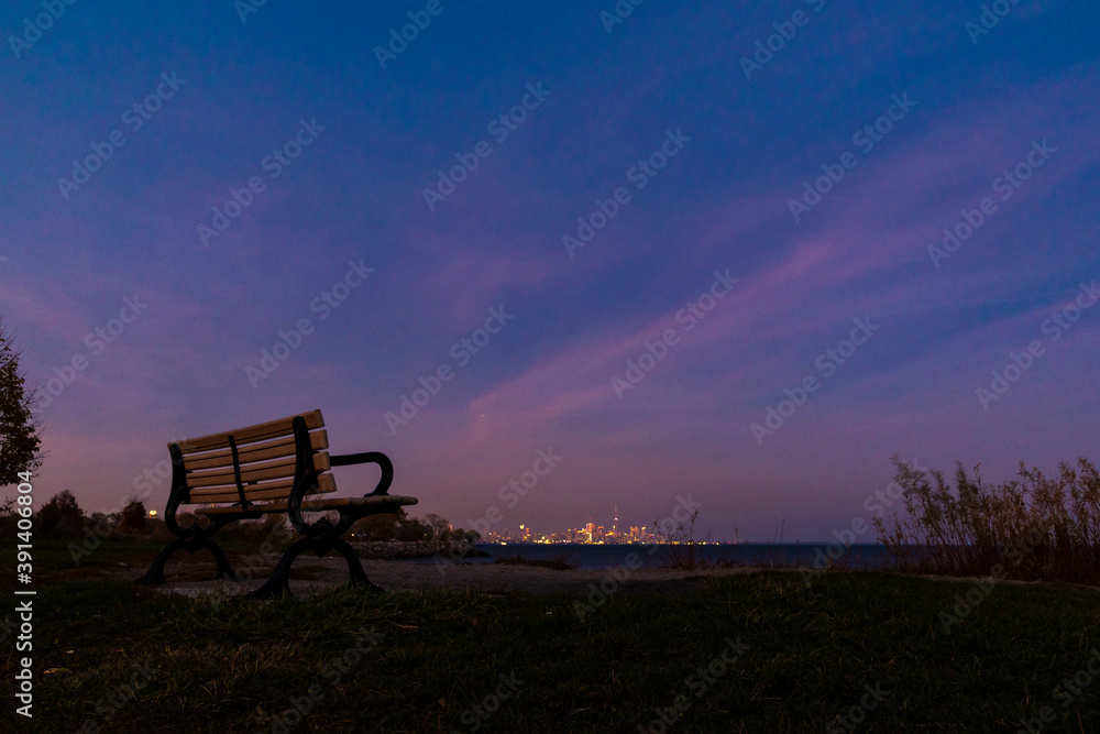 A single empty park bench sits by the side of Lake Ontario, looking across towards the downtown Toronto skyline, in Sam Smith Park in Toronto (Etobicoke), Ontario. A single empty park bench sits by the side of Lake Ontario, looking across towards the downtown Toronto skyline, in Sam Smith Park in Toronto (Etobicoke), Ontario.