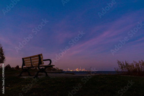 A single empty park bench sits by the side of Lake Ontario, looking across towards the downtown Toronto skyline, in Sam Smith Park in Toronto (Etobicoke), Ontario.