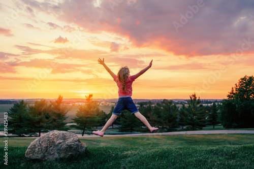 Cute child jumping up with her arms raised on hill at sunset.  Girl kid having fun outdoor enjoying life at sunrise. Local staycation vacation travel. View from back. Happy childhood lifestyle.  © anoushkatoronto