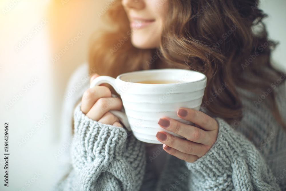 Close-up of female hands with a mug of drink. Beautiful girl in a gray sweater is holding a cup of tea or coffee in the morning sunlight. Mug for your design. High quality photo. Close-up of female hands with a mug of drink. Beautiful girl in a gray sweater is holding a cup of tea or coffee in the morning sunlight. Mug for your design. High quality photo.