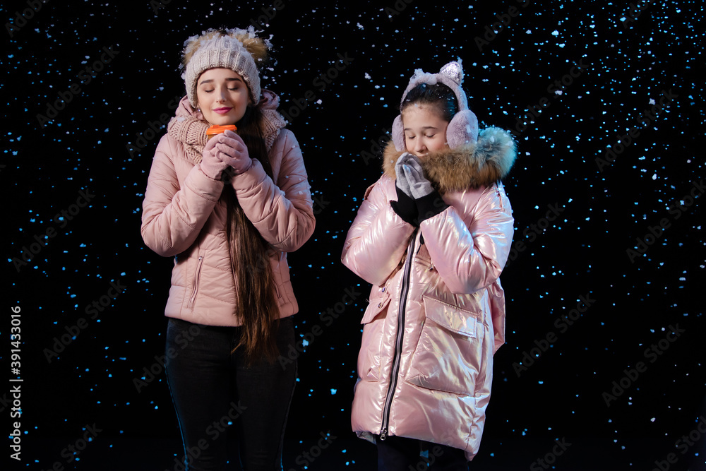 The frozen mother and daughter warm up and enjoy the aroma of the hot drink. Picture taken in a dark studio against a black background during a snowfall. Close up. The frozen mother and daughter warm up and enjoy the aroma of the hot drink. Picture taken in a dark studio against a black background during a snowfall. Close up.
