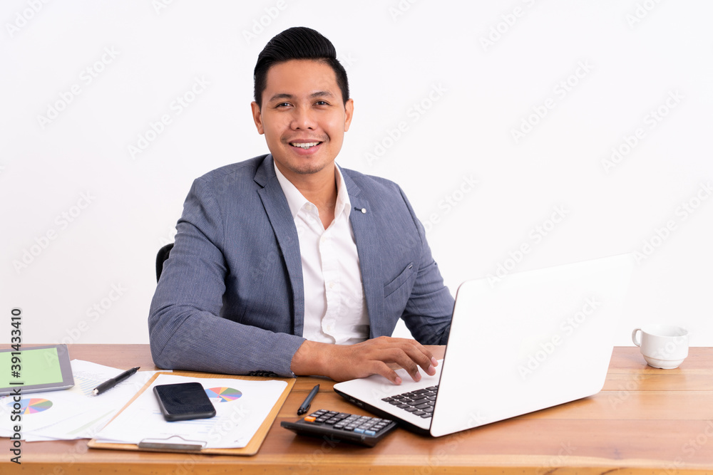 an attractive and cheerful young business man working on a laptop and smiling sitting down. On modern office desk an attractive and cheerful young business man working on a laptop and smiling sitting down. On modern office desk