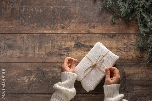 Top view on young woman's hands holding Christmas gift box wrapped in white paper on rustic wooden background. Preparing for holidays and celebration.