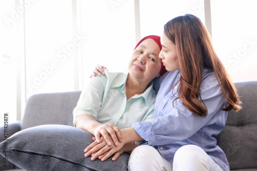 Young Asia woman with her mother wearing a headscarf fighting cancer sits on the couch and her arm wrapped around her mother.