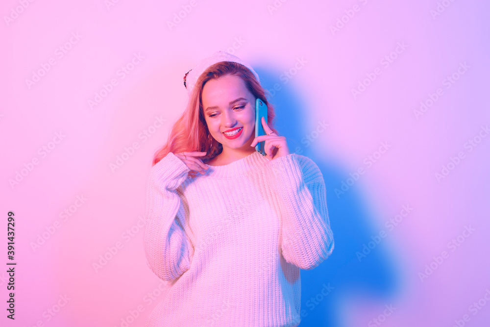 Girl in a white sweater and santa hat in neon light. Woman with professional makeup and red lips. The model speaks on the phone and smiles. New Year Christmas.