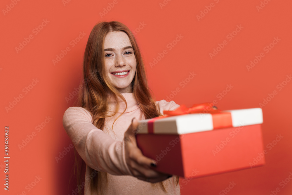 Fototapeta premium Adorable caucasian woman with freckles and red hair is giving at camera a present box smiling on a red studio wall