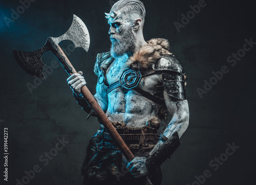 Naked and pale skinned god with horns and blue eyes holding two handed axe and looking at it in dark background.