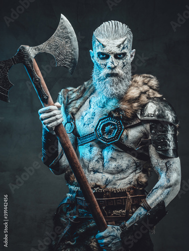 Shirtless and risen from the dead warrior with iced skin and horns in dark armour holding two handed axe in dark background.