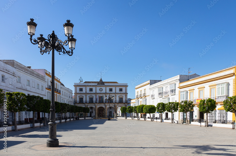The town hall at the end of the historic market square in the historic mountain village of Medina-Sidonia in Andalusia in southern Spain with green trees. The houses are all painted white.