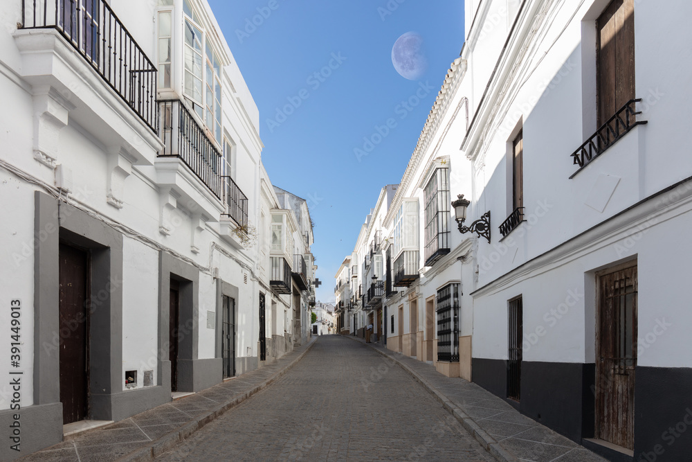 An alley in the historic mountain village of Medina-Sidonia in Andalusia in southern Spain. The houses are all painted white. It's early morning with sunshine. The moon is in the sky.