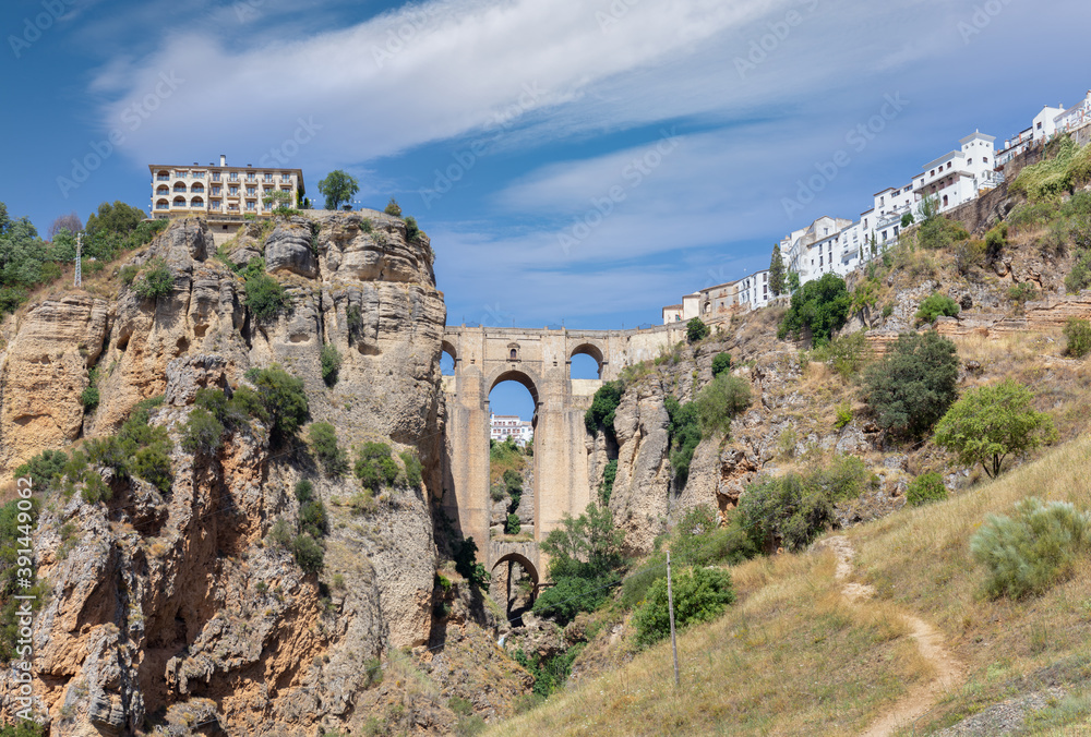 Fototapeta premium The famous arch bridge of Ronda in Andalusia in summer with a blue cloudy sky. The city is on top of the plateau. At the very bottom is a small waterfall.