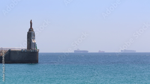 The statue of Christ looks towards the Atlantic from the pier in Tarifa in southern Spain. In the background on the Strait of Gibraltar are large container ships. There is a lot of haze in the air.